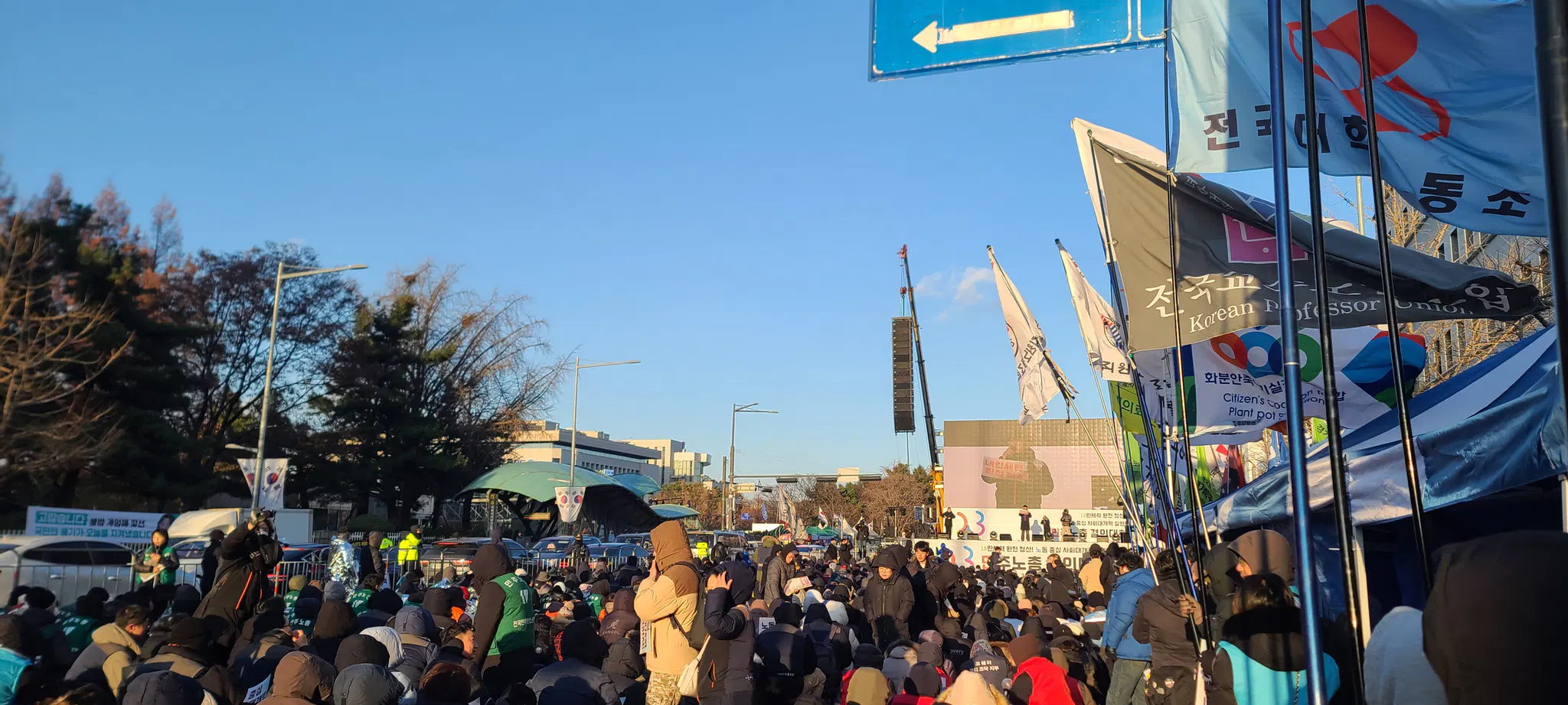 사람들의 앞의 무대를 보고 서 있으며 그 오른쪽에는 여러 깃발들이 휘날리고 있다.